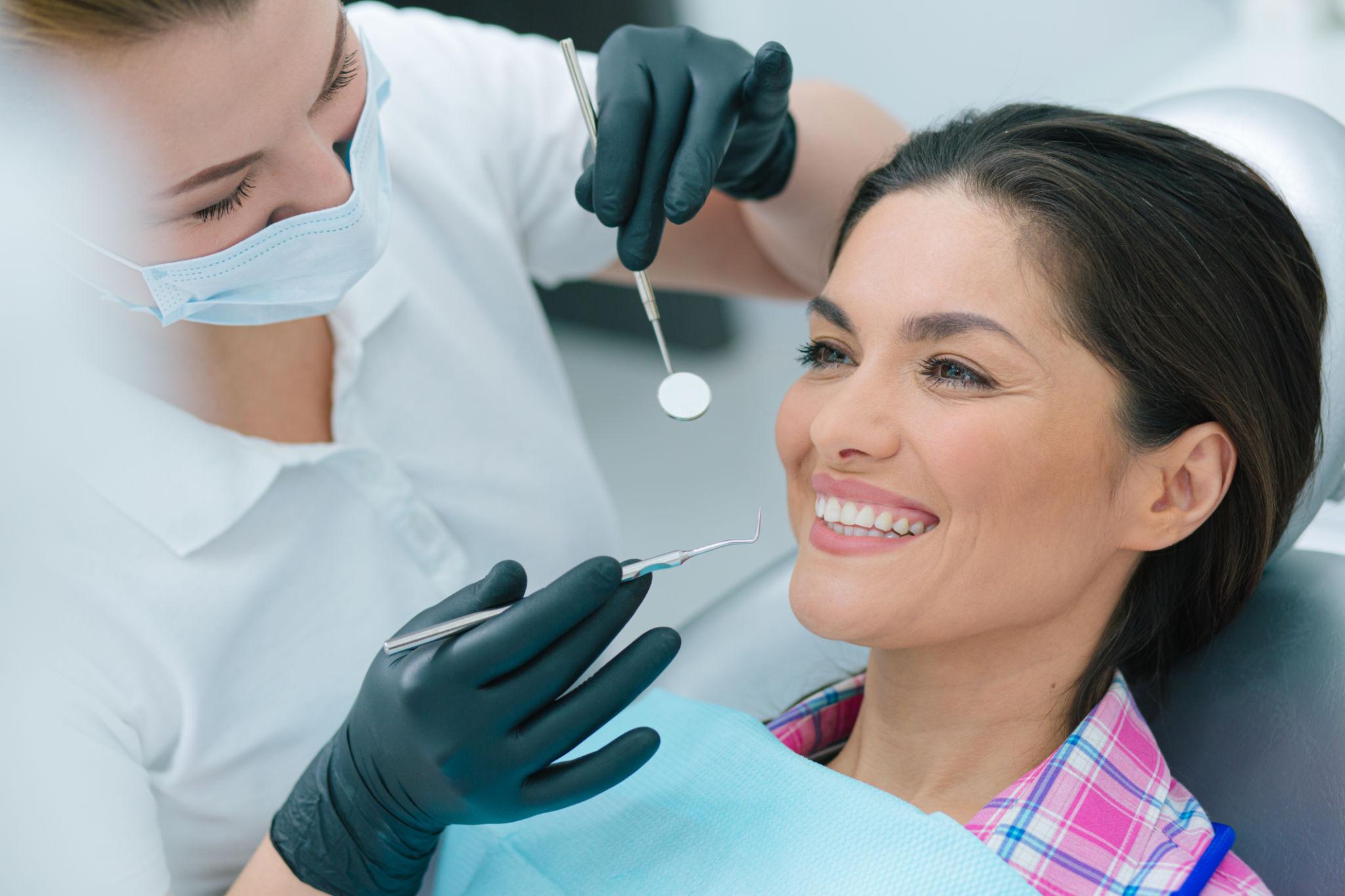 Dentist in a mask and gloves examining a smiling patient during a dental check-up. They hold dental tools near the patient’s mouth.