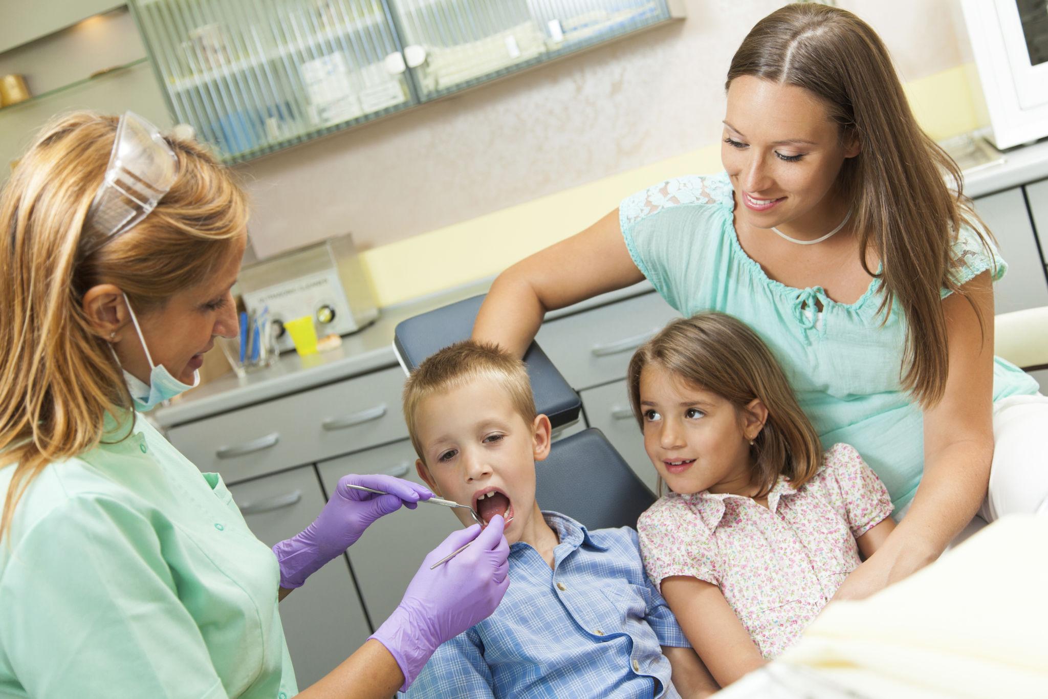 Dentist in green scrubs and purple gloves examining a young boy’s mouth while two children watch beside the chair in a bright clinic.