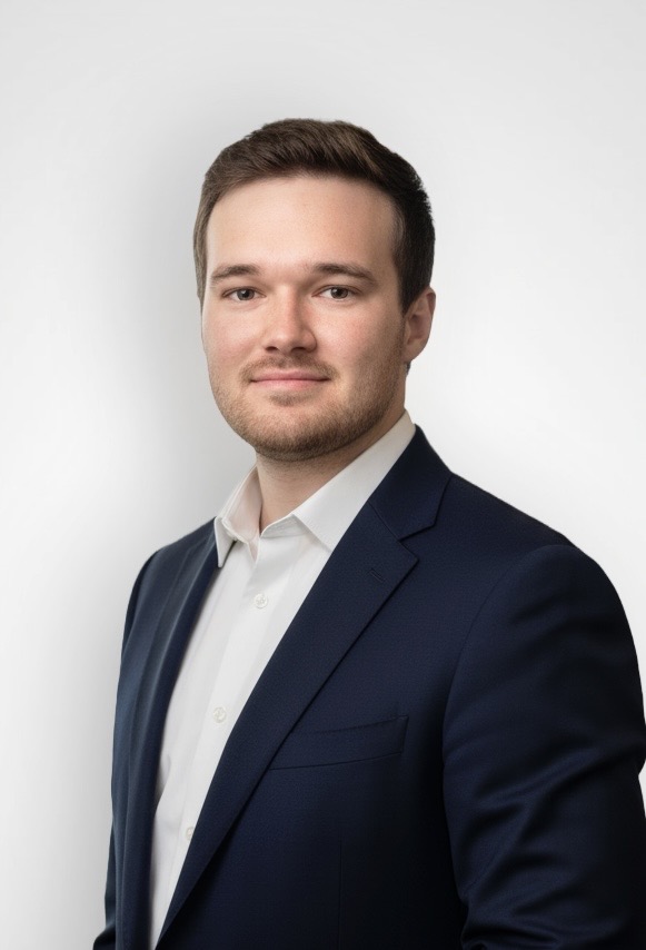 Professional headshot of a man in a navy suit and white shirt against a light neutral background, looking at the camera with a slight smile.