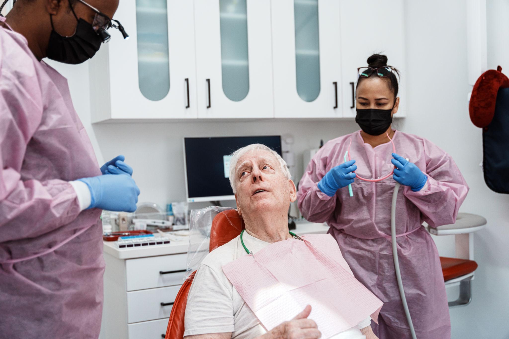 Elderly man seated in a dental chair while two clinicians in pink gowns and masks assist him with dental care.