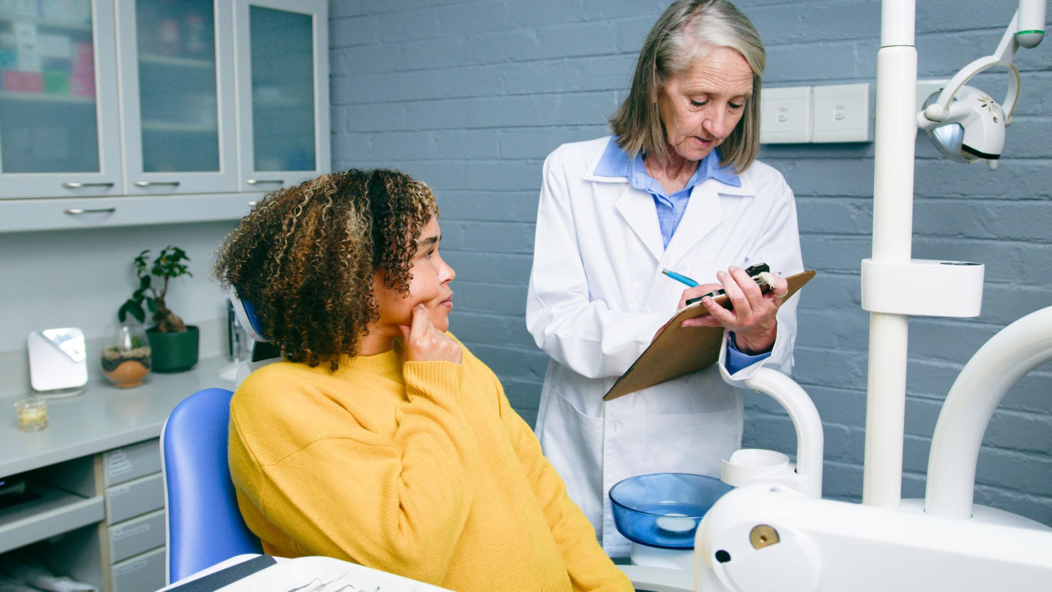 A dentist or nurse in a white coat reviews a clipboard while a patient in a yellow sweater sits in a dental chair, listening attentively.
