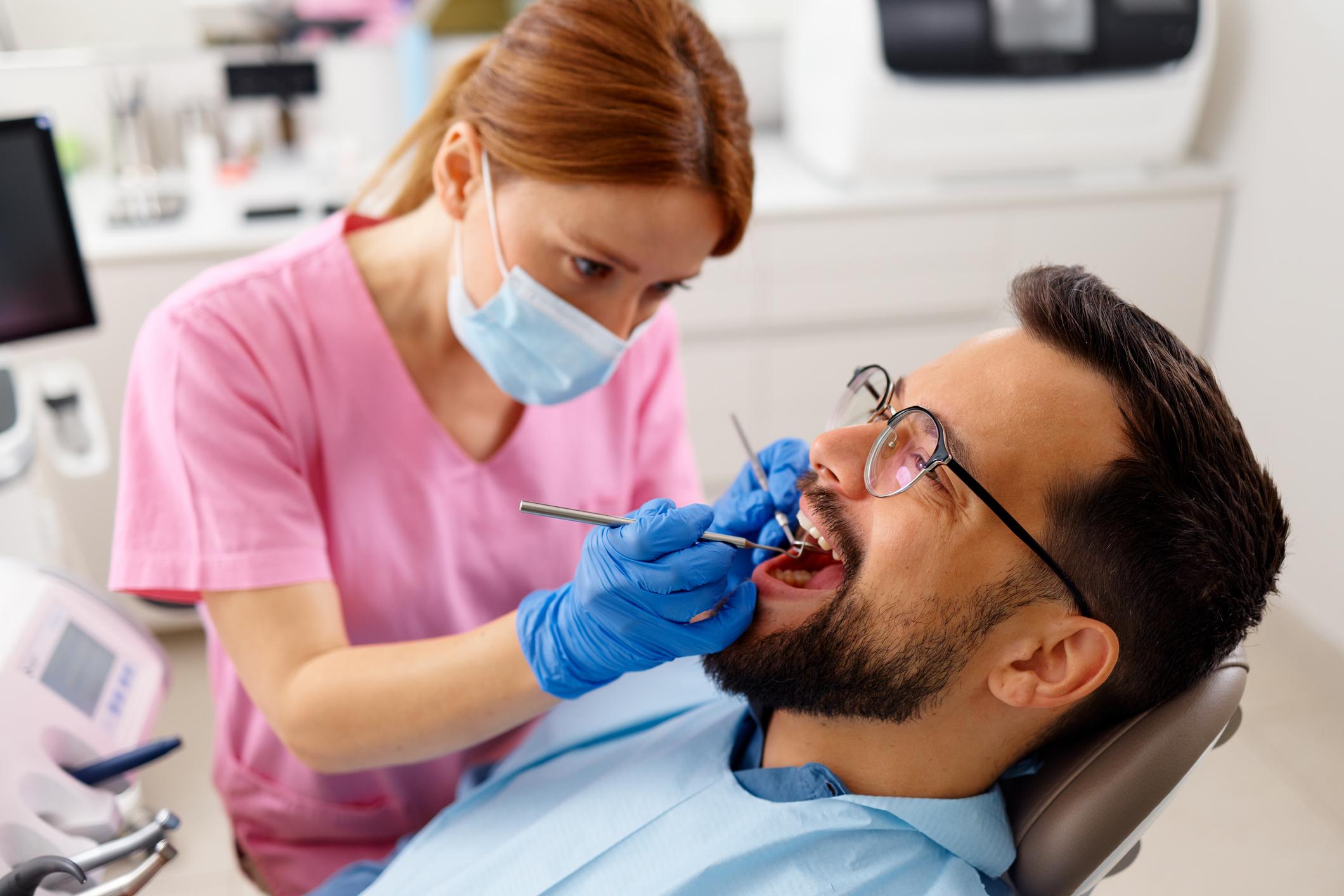 Dentist in pink scrubs and mask treating a male patient wearing glasses in a dental chair, using dental tools in a clinic.