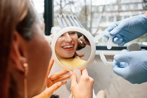 Patient smiling into a small handheld mirror during a dental exam, with a dentist's gloved hands and tools visible in the background.