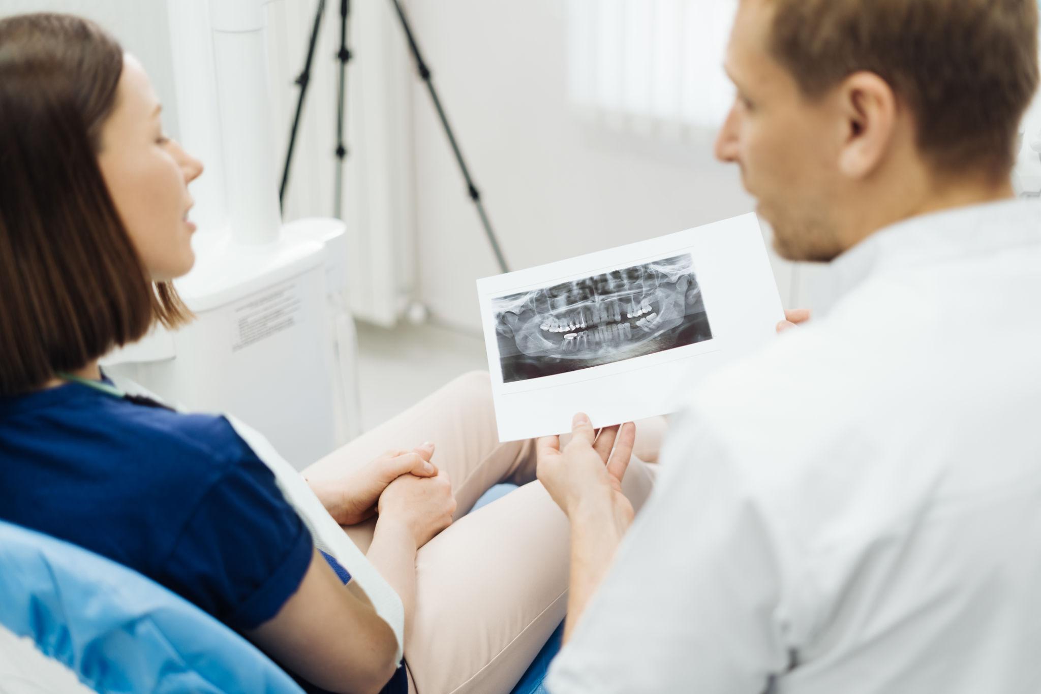 Two clinicians in blue and white coats review a dental X-ray film together in a clinic, discussing a patient’s teeth