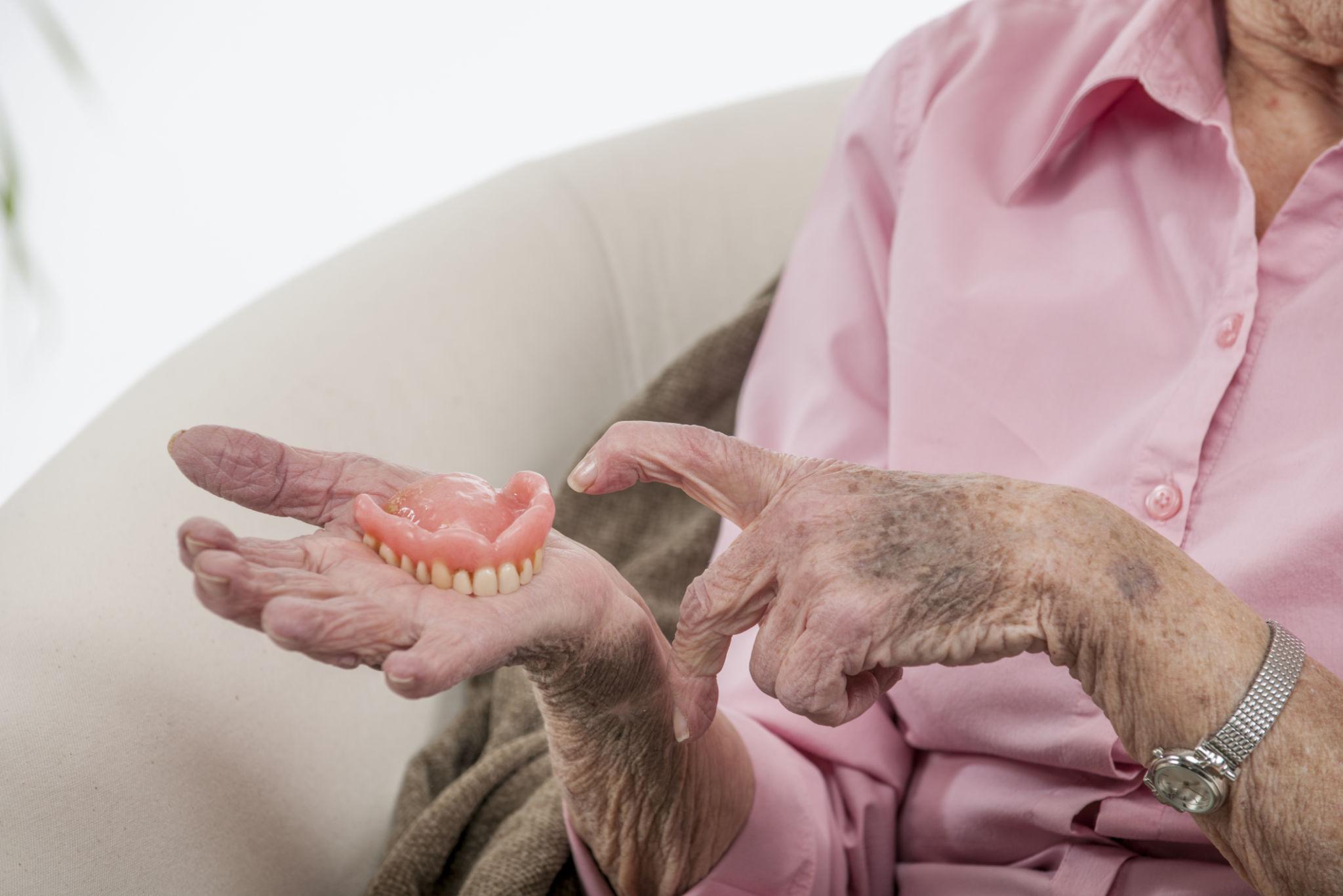 Elderly person in a pink shirt holds a set of dentures in their hands.