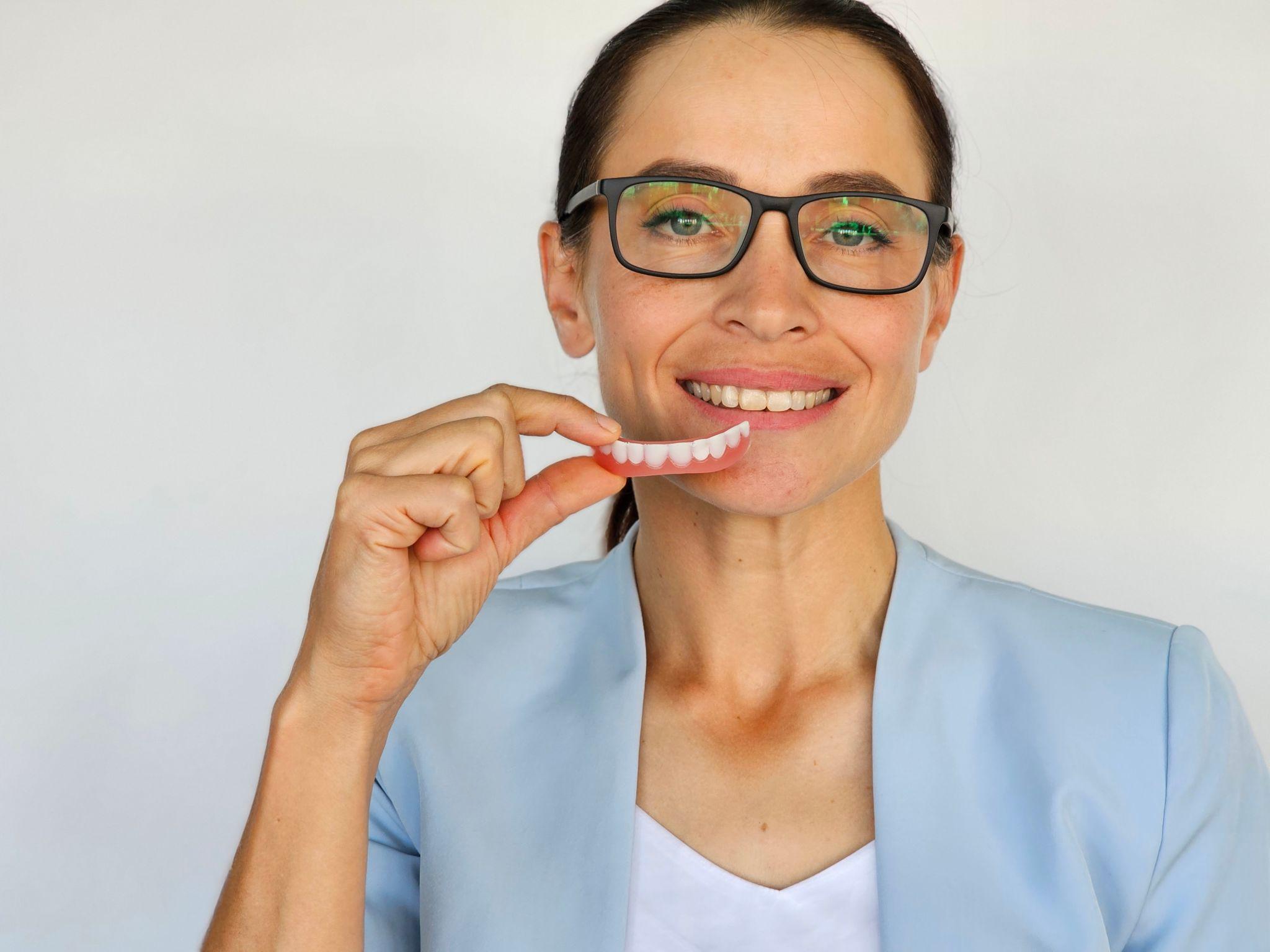 Smiling woman wearing glasses holds a pink dental denture up to her mouth.