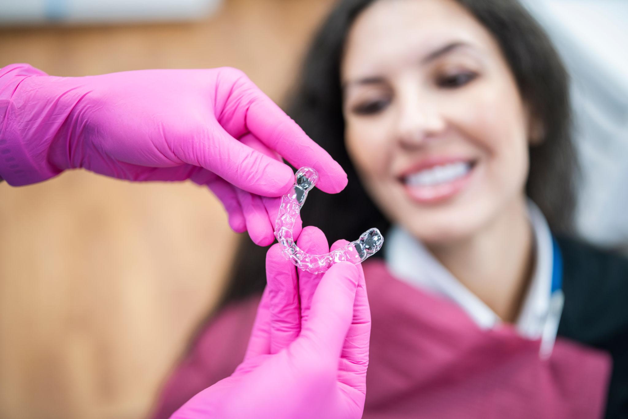 Dental professional in pink gloves presenting a clear dental retainer to a smiling patient in the background