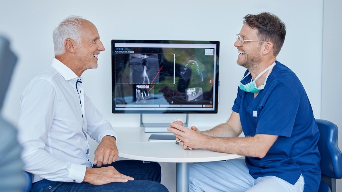 Doctor in blue scrubs and an older patient in a white shirt smile while reviewing medical images on a monitor during a clinic consultation.
