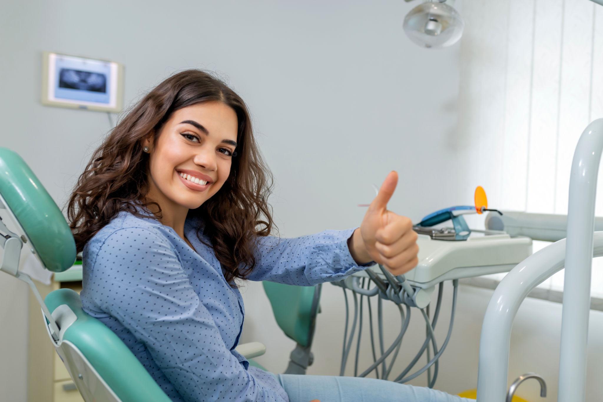 Smiling woman in a dental chair giving a thumbs up, dental equipment visible in the background.