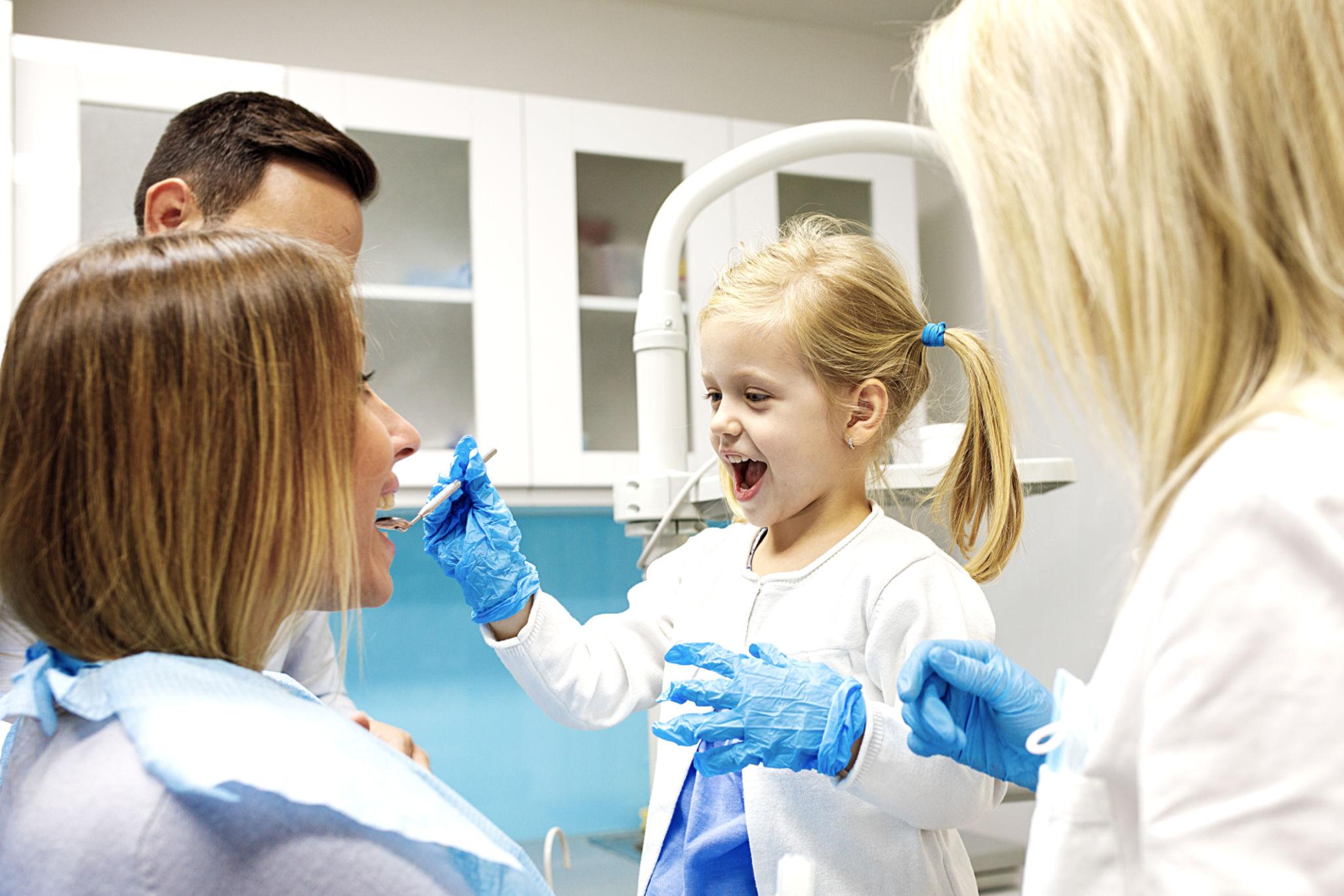 Young girl at a dental checkup, smiling as a clinician uses tools near her mouth with blue gloves.
