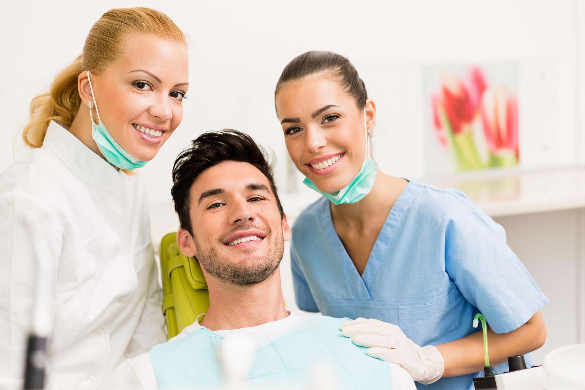 Smiling patient in a dental chair taking a selfie with two dental professionals in scrubs and masks nearby in a clinic.