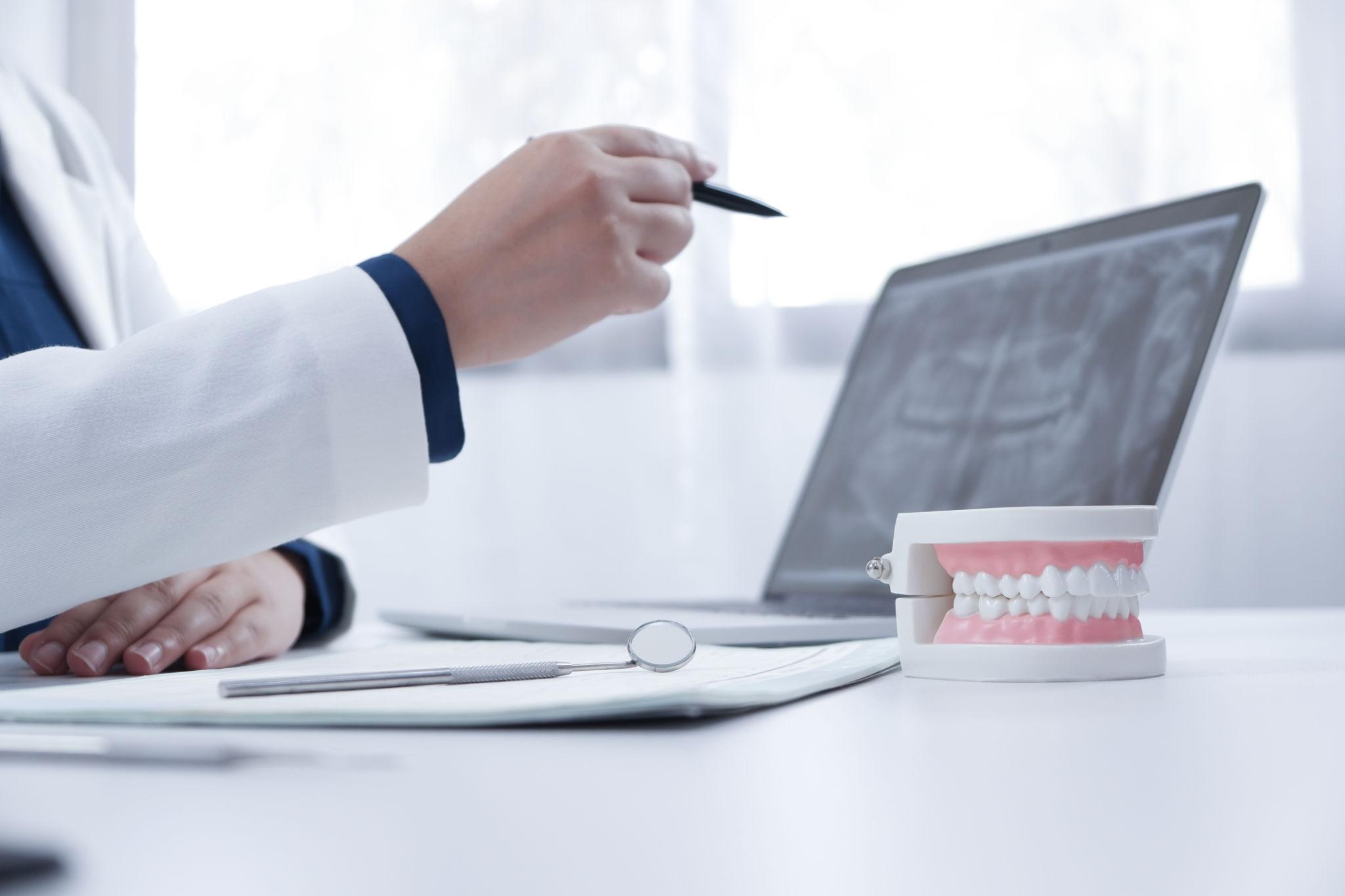 Dentist in a white coat writing with a pen beside a laptop and a dental model on the desk.