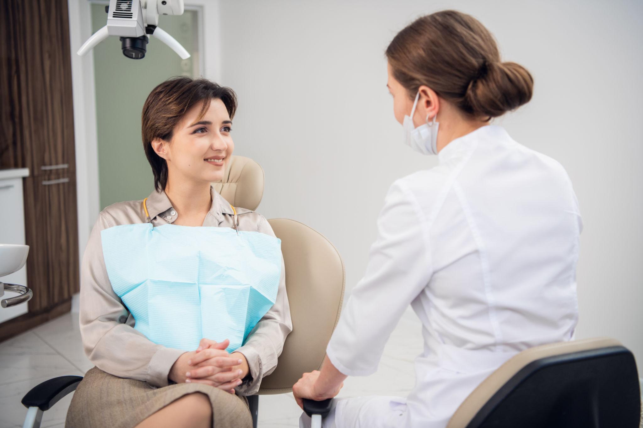 A smiling patient sitting in a dental chair with a blue bib, talking to a dentist wearing a mask in a clinic.