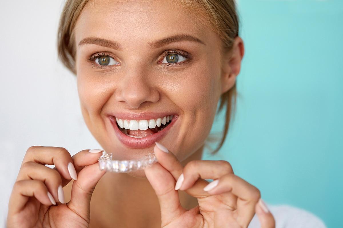 Happy woman showing a clear aligner between her fingers, smiling widely with bright teeth against a turquoise background.