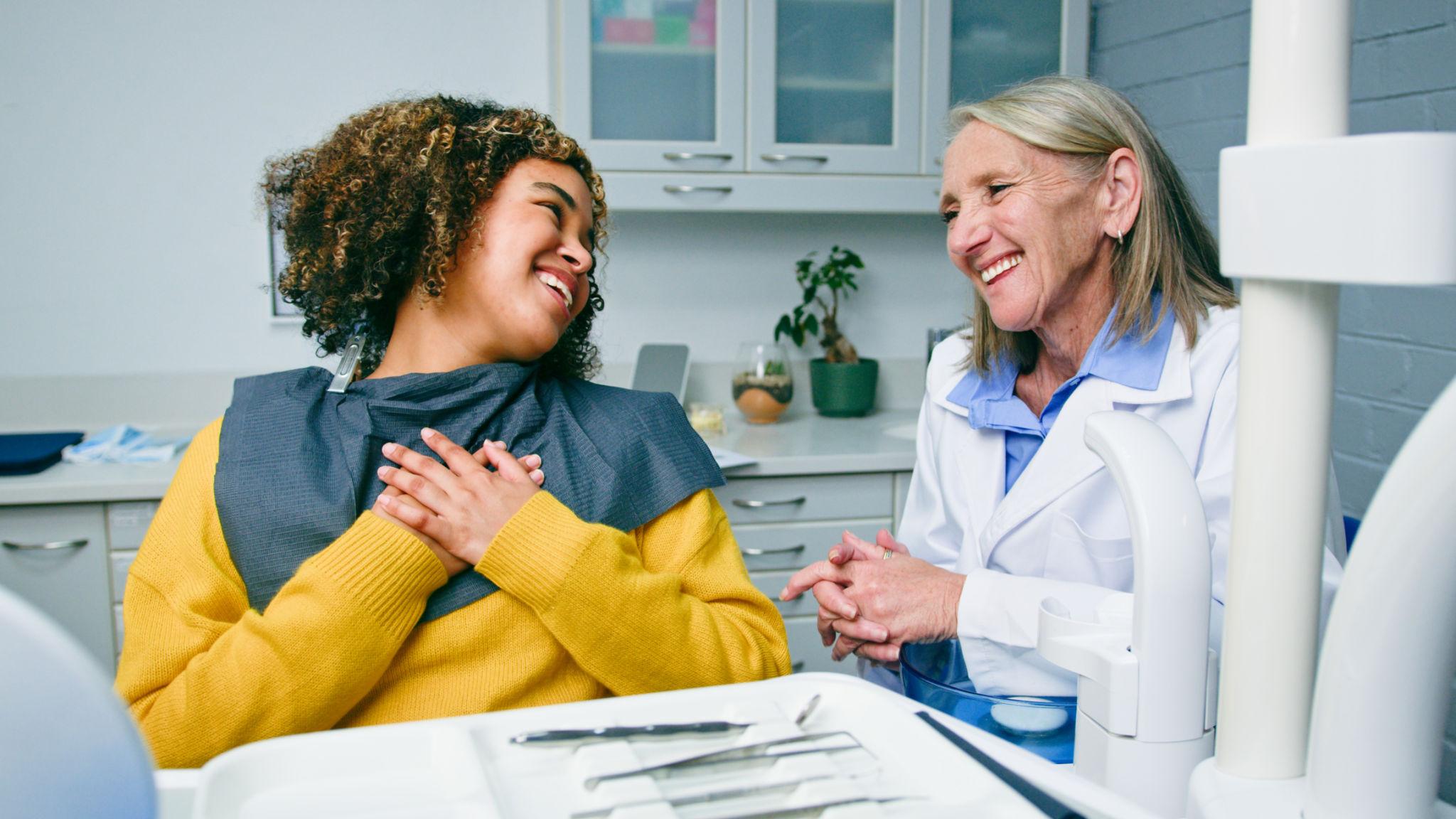 Patient and dentist share a smile during a dental visit in a clinic, with the patient wearing a bib and a yellow sweater.