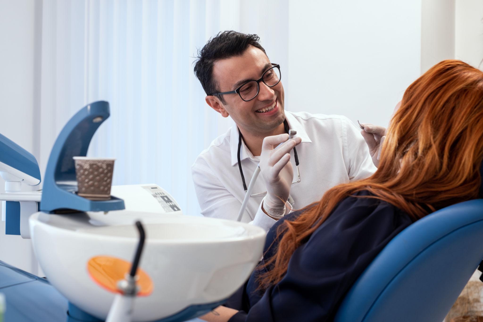 Dentist in a white coat wearing gloves examines a patient in a dental chair, smiling.