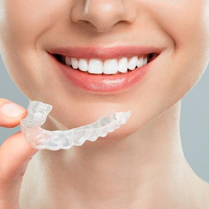 Close-up of a smiling person holding a clear dental aligner between their fingers near the lower teeth.