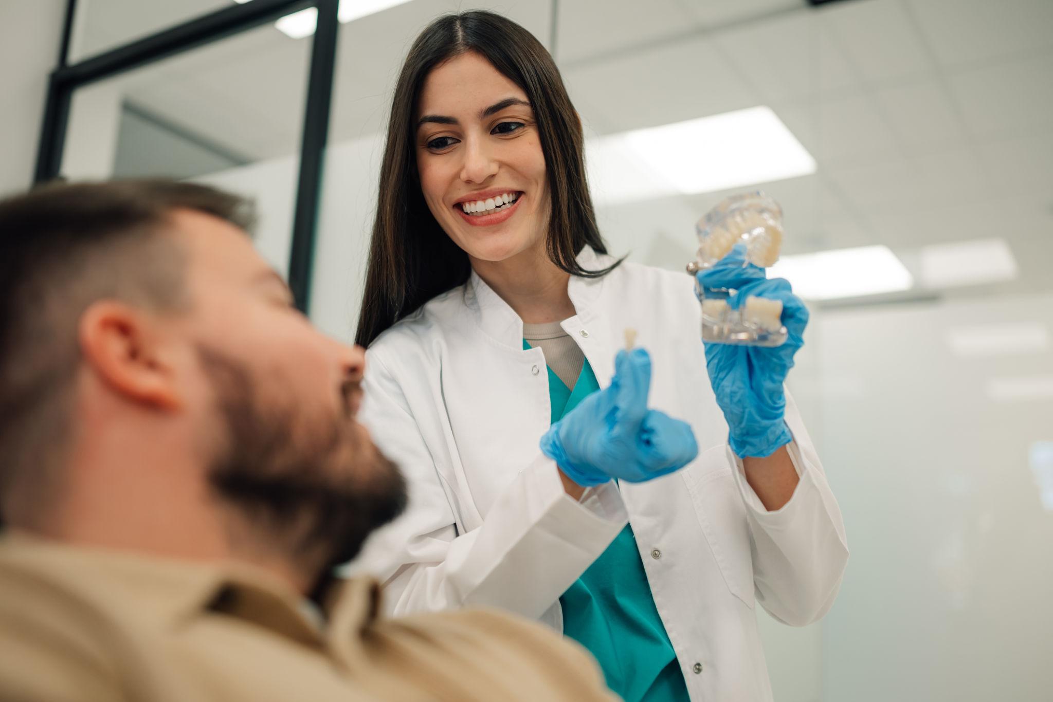 Female dentist in a white coat showing a dental model to a patient, smiling in a bright clinic.