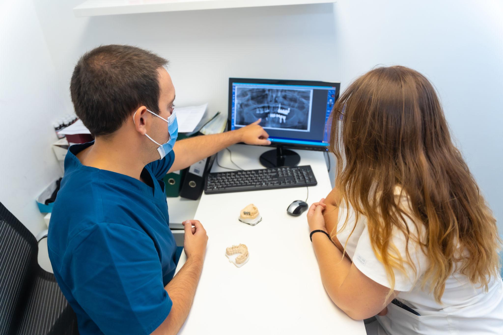 Dentist in blue scrubs points to a dental X‑ray on a computer monitor for a patient in a clinic.