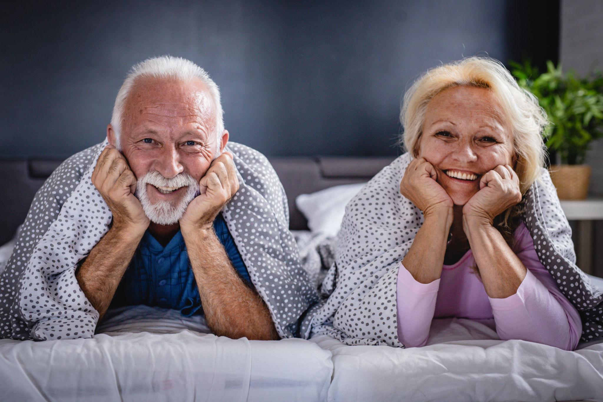 Smiling elderly couple lying in bed with their hands under their chins, wrapped in polka-dot blankets in a cozy bedroom scene.