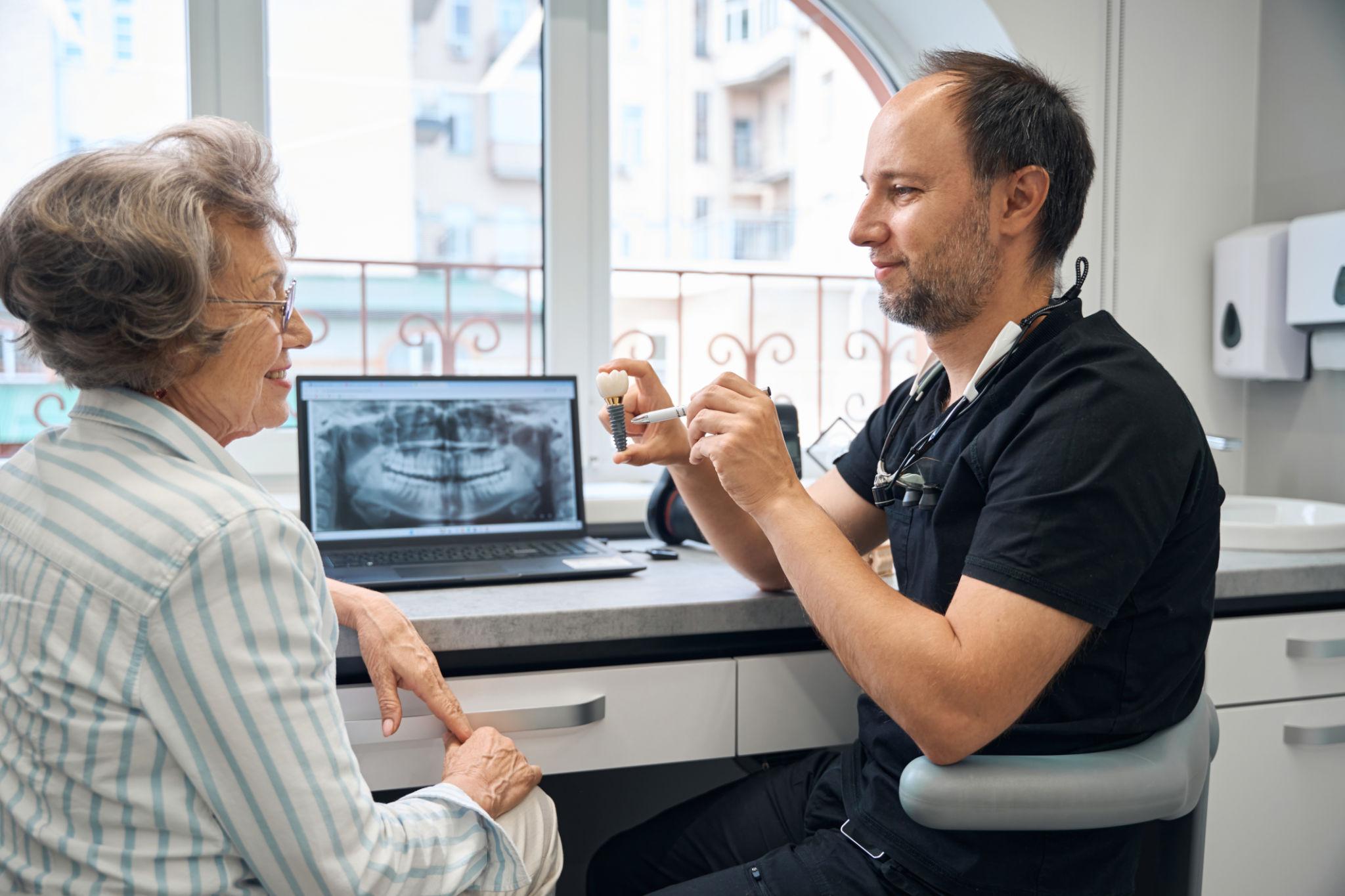 Dentist demonstrating a tooth model to an elderly patient, with an X-ray displayed on a laptop screen.