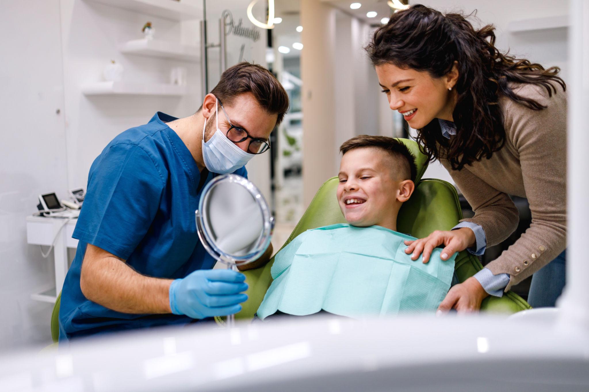 Dentist in blue scrubs and mask examining a smiling boy in a dental chair as his mother leans in nearby.