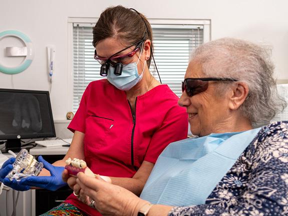Dental assistant in red scrubs, mask, and loupes shows a dental model to an elderly patient wearing protective glasses and a bib in a clinic.