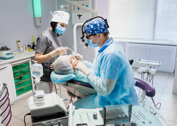 Dentist and assistant wearing masks perform a dental procedure on a patient in a chair surrounded by dental equipment.
