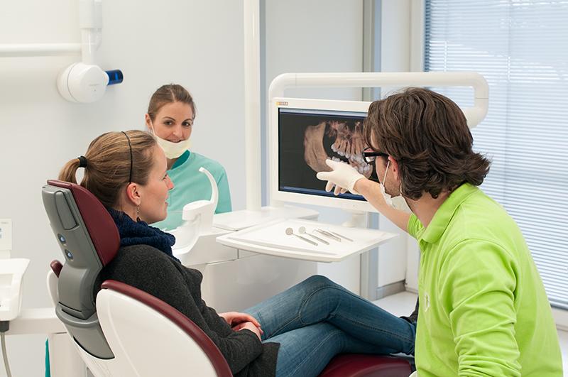 Dentist in a green shirt points to a dental scan on a monitor as a patient sits in a dental chair, with an assistant nearby in a clinic.